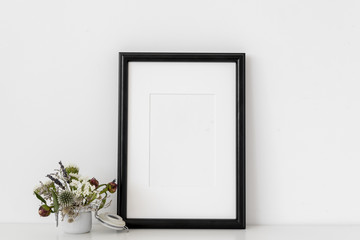 Black a4 portrait frame mockup with dried field flowers in small white pot on white wall background. Empty frame, poster mock up for presentation design. Template frame for text, lettering, modern art