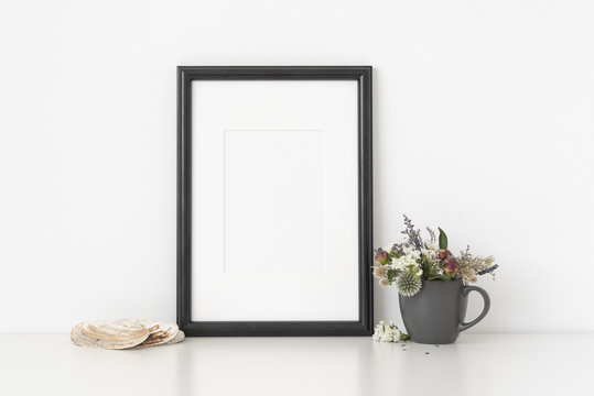 Black a4 portrait frame mockup with dried field wild flowers in vase and seashells on white wall background. Empty frame, poster mock up for presentation design.