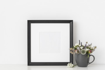 Black square portrait frame mockup with dried flowers in gray mug on white wall background. Empty...
