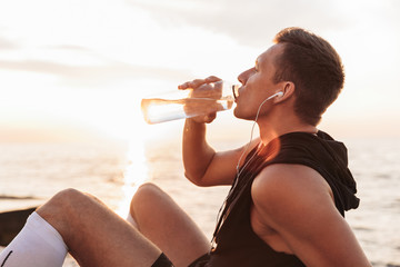 Sportsman outdoors at the beach listening music with earphones drinking water.