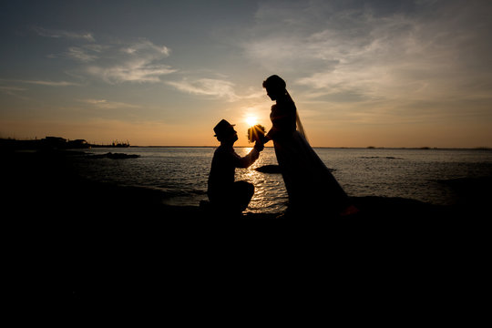 A silhouette of a young man, down on one knee and holding a bouquet, proposing to his girlfriend.will you marry me images.Young couple in love at beautiful sunset.
