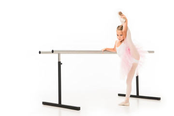 little ballerina in tutu stretching leg at ballet barre stand isolated on white background © LIGHTFIELD STUDIOS