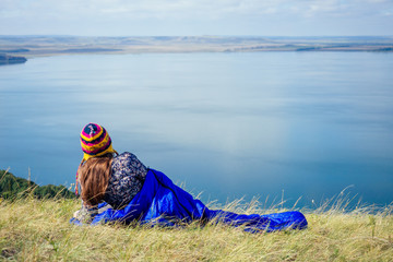 tourist woman hiker wrapped up in a blue sleeping bag on a grass lake background mountains