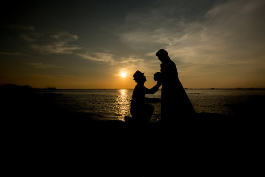 A silhouette of a young man, down on one knee and holding a bouquet, proposing to his girlfriend.will you marry me images.Young couple in love at beautiful sunset.