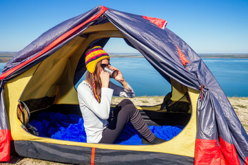 Young woman in a funny hat from Nepal sitting in a blue sleeping bag in the tent holding a thermos...