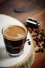 A cup of coffee with small white ceramic dish full of coffee beans on wooden background.