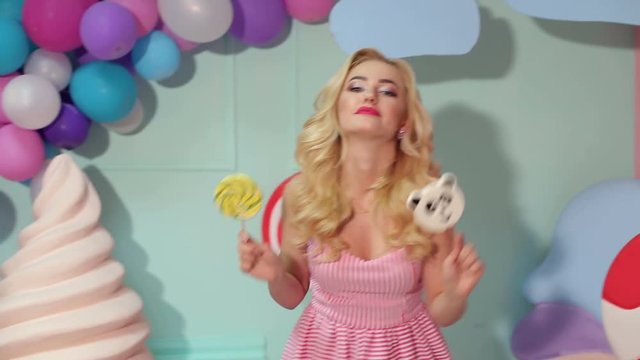 A cheerful girl with big lollipops in her hands is dancing and having fun in a bright room with balloons and a large plastic ice cream, she is dressed like a barbie. Barbie girl. Candy girl.