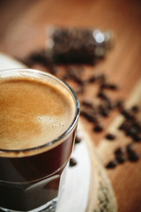 A cup of coffee with small white ceramic dish full of coffee beans on wooden background.