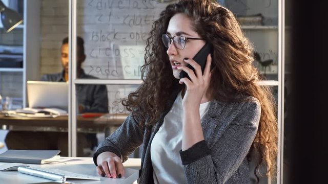 Young intelligent businesswoman in eyeglasses talking on cell phone while working late in office; black businessman using laptop behind glass wall