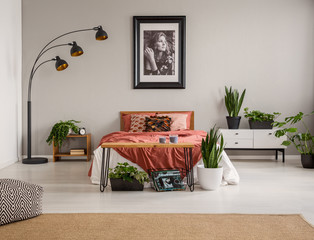 Pouf on carpet and plants in front of red bed in grey bedroom interior with poster and lamp. Real photo