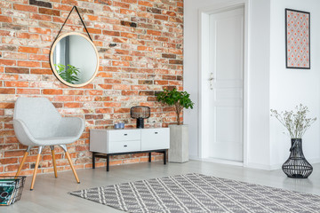 Grey armchair and cupboard in apartment interior with carpet and mirror on red brick wall. Real...