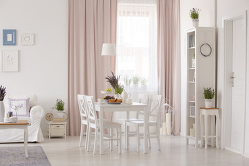 White chairs at table near couch in pink apartment interior with drapes and plants. Real photo