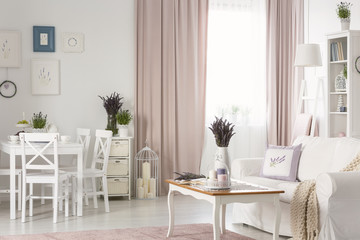 White chairs at dining table near posters in flat interior with pink drapes and settee. Real photo