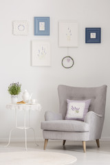 Flowers on table next to grey armchair with cushion in white flat interior with posters. Real photo