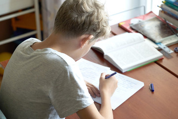 A cacausian blonde teenager boy with bad posture sitting at a table and writing in a copybook. Slouching blonde teenager boy doing school homework. - obrazy, fototapety, plakaty