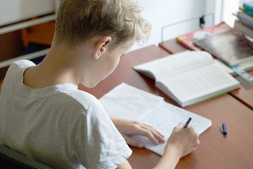  Blonde teenager boy doing school homework sitting at the table.