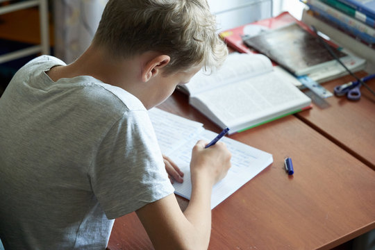 A cacausian blonde teenager boy with bad posture sitting at a table and writing in a copybook. Slouching blonde teenager boy doing school homework.
