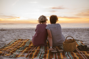 Young lesbian couple watching a romantic sunset at the beach - obrazy, fototapety, plakaty