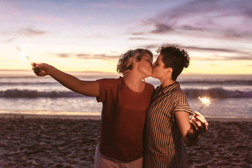 Romantic lesbian couple kissing and holding sparklers at the beach