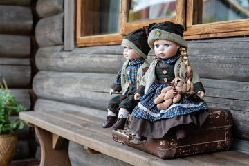 Ancient porcelain dolls sits on suitcase near log farmhouse.