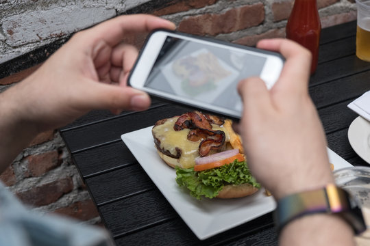 Man takes photo of food with mobile phone at an outdoor bar.  Taking a picture of your food with your phone. Hamburger, fries, and beer on a white plate outside on a black table focus on food.