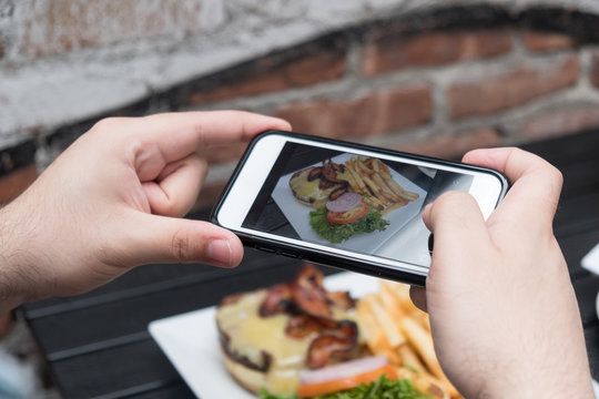 Man takes photo of food with mobile phone at an outdoor bar.  Taking a picture of your food with your phone. Hamburger, fries, and beer on a white plate outside on a black table, point of view shot.