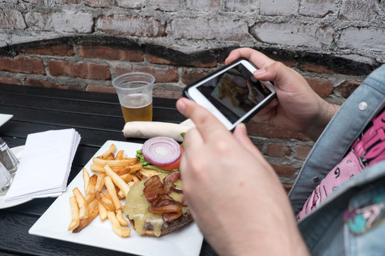 Man holding a mobile phone taking a photo of his food. Smartphone food photography. Taking a picture of hamburger, beer, and french fries at an outdoor bar with a mobile phone.