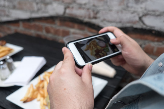 Man holding a mobile phone taking a photo of his food. Smartphone food photography. Taking a picture of hamburger and french fries at an outdoor bar with a smartphone.