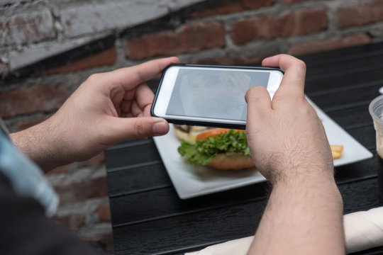 Man holding a mobile phone taking a photo of his food. Smartphone food photography. Taking a picture of hamburger and french fries at an outdoor pub.