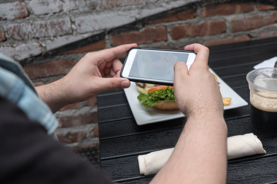 Man takes a photo of his dinner with his cell phone. Mobile phone photography. Taking a photo with phone of hamburger, french french fries and dark beer on white plate on black table outside.
