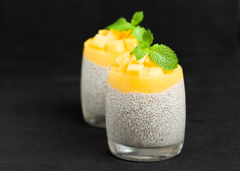 Chia seeds pudding with mango puree in glasses with mint leaves and cut ripe tropical fruit on black background.