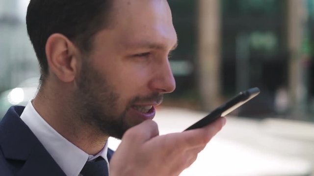 Hansome young caucasian businesman with fair hair and beard wearing a dark suit and tie talking on smartphone and smiling on a summer day. Tilt up slow motion medium shot