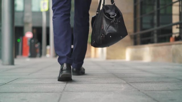 Legs of unrecognizable businessman walking in a summer street. He is wearing a suit and carrying a leather bag walking from the camera. Tracking slow motion low medium shot