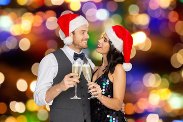 celebration, people and holidays concept - happy couple in santa hats with glasses of non alcoholic champagne at christmas or new year party over festive lights background