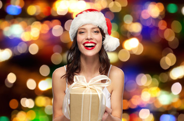 christmas, holidays and people concept - happy smiling young woman with red lipstick in santa hat holding present over festive lights background