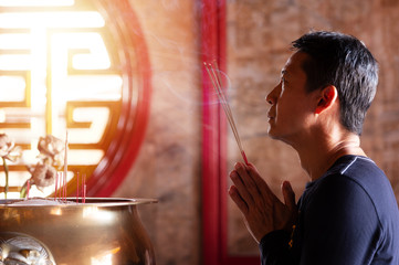 Faith and religious. .Man hands holding joss sticks blessing in front of altar table at chinese shrine with sunlight through window,side view..