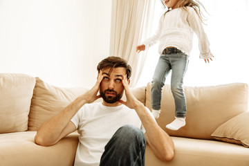 Parenthood. Family. Tired. Dad is having a headache while his little daughter is jumping on the...