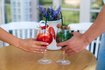 A curious plan cropped frame of a man and a woman take glasses with bright tasty berry cocktails. Two glasses and a decorative white vase with flowers stand on a table on a light background.