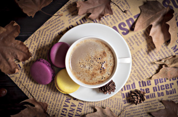 Autumn, fall dried leaves, hot steaming cup of cappuccino coffees, macaroons, cones, newspaper on wooden table background. Seasonal, evening coffee, Sunday relaxing and still life concept. Flat lay. 