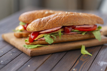  Two baguette sandwiches with salami, cheese, arugula salad, tomatoes and onion on a cutting board....