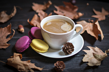 Autumn, fall dried leaves, hot steaming cup of cappuccino coffees, macaroons, cones, newspaper on wooden table background. Seasonal, evening coffee, Sunday relaxing and still life concept. Flat lay. 