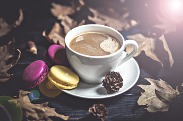 Autumn, fall dried leaves, hot steaming cup of cappuccino coffees, macaroons, cones, newspaper on wooden table background. Seasonal, evening coffee, Sunday relaxing and still life concept. Flat lay. 