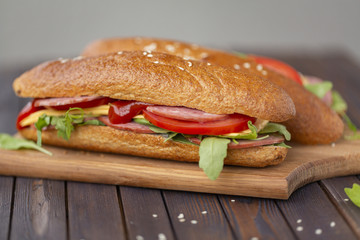  Two baguette sandwiches with salami, cheese, arugula salad, tomatoes and onion on a cutting board. Sandwiches on dark wooden background. Tasty snack.