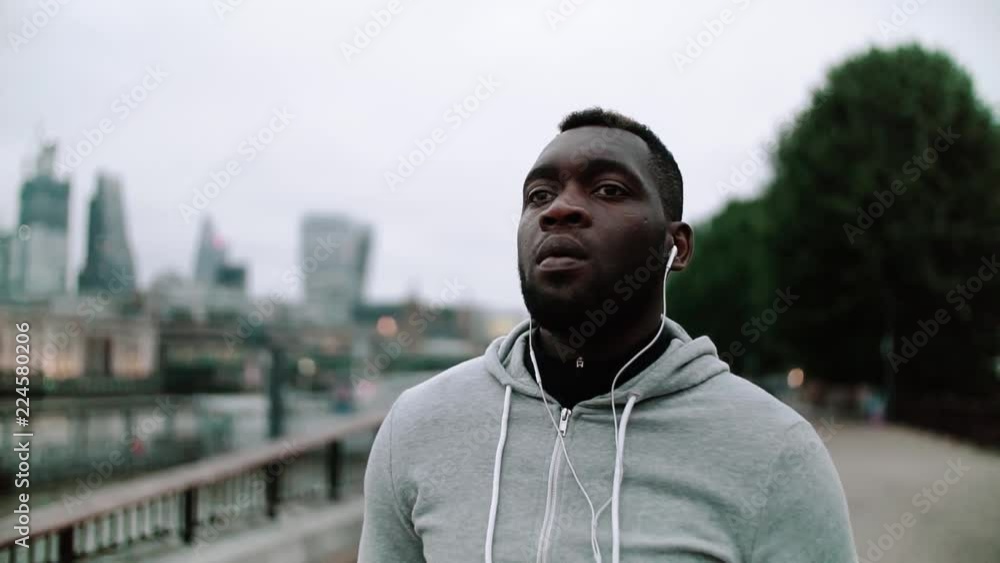 Poster Young sporty black man runner with earphones walking on the bridge in a city.