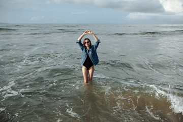 Happy smile brunette Woman wearing black swimsuit and denim jacket at ocean background enjoy walking in ocean, hands raised up