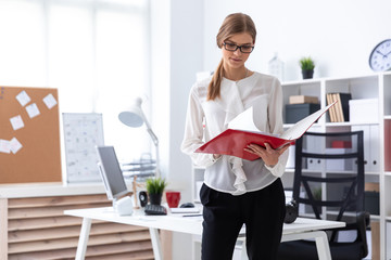 A young girl stands near a computer desk and scrolls through a folder with documents.
