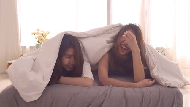 Beautiful happy Asian lesbian couple or friends wear pajamas wake up kissing each other have romantic time on bed in bedroom at home. Lifestyle lesbian LGBT couple relax at home concept.