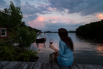 Young brunette woman sitting on a lakeside dock at sunset enjoying a glass of red wine.