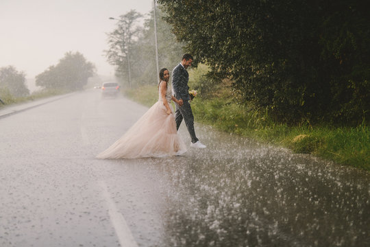 Silly young just married couple crossing road on rainy day. Walking in wet ceremonial clothes.