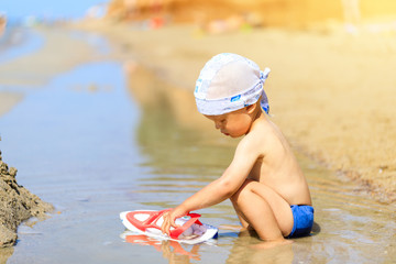 Baby boy sitting on the beach near the water and plays with a toy ship